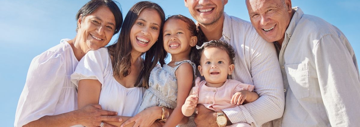 Multi-generational family in white clothing smiling together outdoors against blue sky.