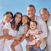 Multi-generational family in white clothing smiling together outdoors against blue sky.