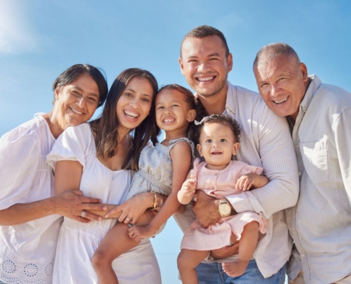 Multi-generational family in white clothing smiling together outdoors against blue sky.
