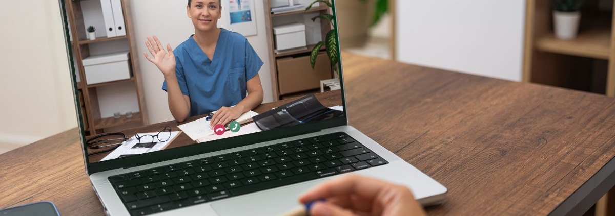 Person video calling with healthcare provider on laptop at wooden desk in home office setting.
