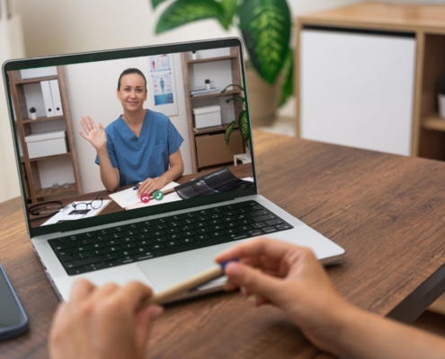 Person video calling with healthcare provider on laptop at wooden desk in home office setting.