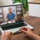 Person video calling with healthcare provider on laptop at wooden desk in home office setting.