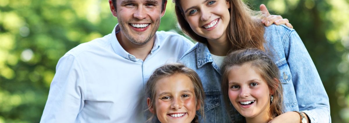 Happy family of four smiling outdoors in bright sunlight with green foliage background.
