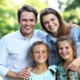 Happy family of four smiling outdoors in bright sunlight with green foliage background.