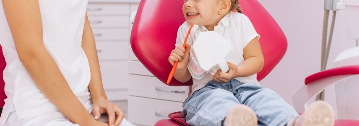 Young child sitting in pink dental chair holding toothbrush, smiling at dentist in Jacksonville, NC.