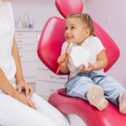 Young child sitting in pink dental chair holding toothbrush, smiling at dentist in Jacksonville, NC.