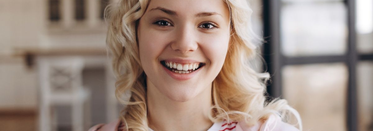 Young woman with blonde wavy hair smiling brightly, wearing a red and white striped shirt and pink cardigan.